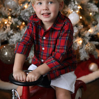 Child in a red plaid shirt sitting on a toy fire truck in front of a decorated Christmas tree.