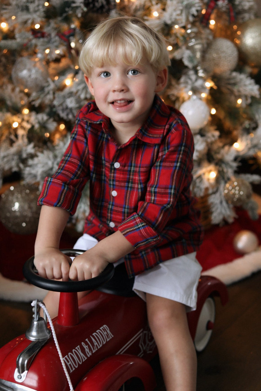 Child in a red plaid shirt sitting on a toy fire truck in front of a decorated Christmas tree.